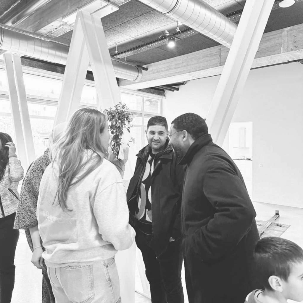 Een groep mensen staat gezellig in gesprek in een modern, industrieel ingericht gebouw met grote ramen en zichtbare luchtkanalen. De sfeer is informeel en vriendelijk. Op de voorgrond is een kind deels zichtbaar. De foto is in zwart-wit.