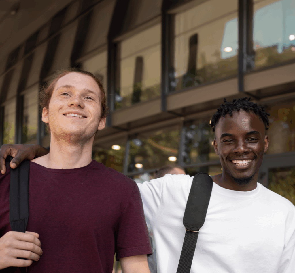 Twee jonge mannen staan lachend naast elkaar met een arm om elkaars schouder, voor een modern school- of kantoorgebouw. Ze dragen casual kleding en rugtassen, wat wijst op een vriendschappelijke en informele setting, mogelijk studenten.